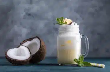 pina colada smoothie in a glass mug with a slice of pineapple on top and a sliced coconut next to it