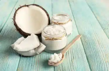 coconut oil in two glass jars with a wooden spoon with more coconut oil in front, and coconut chunks and a half coconut next to them