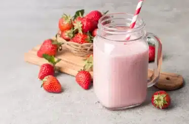 Homemade strawberry milk in a glass mug with a striped straw in it and a heaping pile of strawberries behind in a small woven basket, plus more strawberries scattered around