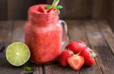 red homemade slushie with strawberries scattered around and a cut lime