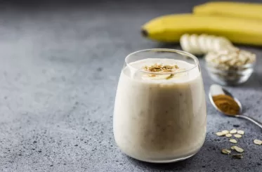 Maca powder smoothie in a glass with a spoonful of maca powder, a small glass bowl of oats, and two bananas in the background
