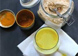 A cup of golden milk latte (turmeric milk) on a napkin surrounded by turmeric and a jar filled with powder mix.