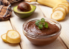A bowl of chocolate avocado pudding encircled by a sliced banana and an avocado sliced in half.