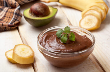 A bowl of chocolate avocado pudding encircled by a sliced banana and an avocado sliced in half.