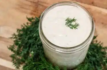homemade ranch dressing in a small glass jar with dill on top and more scattered around, all on a wooden background