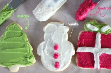 Three decorated Christmas cookies - one green tree labelled "matcha", one white snowman labelled "vanilla", and one pinkish red present labelled "beetroot" underneath three knives with the different colored royal frosting.