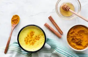 mug of golden milk with turmeric swirled in it; spoon of turmeric next to it, two cinnamon sticks, a small clear bowl of honey, and another bowl of turmeric powder