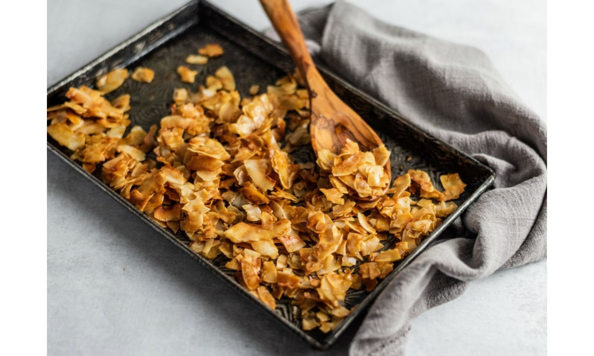 A wooden spoon showing crispy, browned coconut "bacon" on a baking tray on top of a grey tea towel.