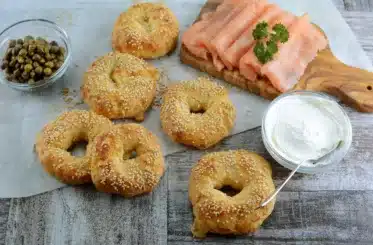Homemade keto bagels on a table surrounded by cream cheese, a wooden paddle topped with sliced salmon, and a bowl of capers