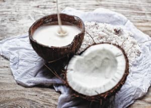 Coconut milk poured into a coconut half-shell next to a half shell showing coconut meat. Next to shredded coconut meat. All on a cloth on a wooden table.