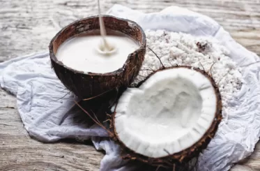 Coconut milk poured into a coconut half-shell next to a half shell showing coconut meat. Next to shredded coconut meat. All on a cloth on a wooden table.