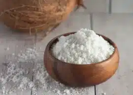 Coconut milk powder in a wooden bowl with coconut milk powder scattered all around and a whole hair coconut in the background