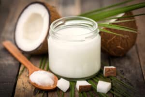 Solidified white coconut oil in a jar with a wooden spoon next to it, pieces of coconut in foreground, and a sliced coconut in the background, all on a wooden table