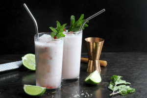Two coconut mojitos (muddled pink and white liquid with a sprig of mint in the top and a straw in each) with a copper jigger and limes