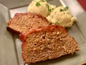 Two juicy slices of Easy Ketchup Meatloaf on a green plate with two mounds of homemade mashed potatoes.