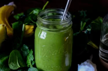 green smoothie with coconut oil in a glass jar with a reusable straw in it; surrounded by green leaves and a white flower