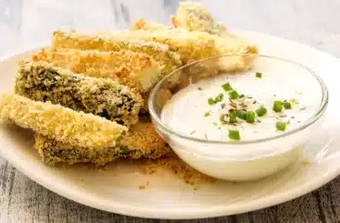Zucchini fries on a plate with a white sauce in a clear bowl next to them