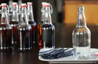 several clear bottles of vanilla extract, with several vanilla pods and an empty bottle on a white dish next to them