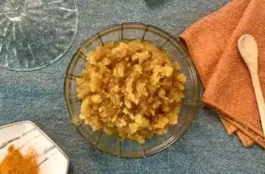 Homemade Turmeric Face Scrub in a glass jar with a lid, an orange cloth, a wooden spoon, and a marble plate of ground turmeric.