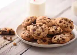 A plate of chocolate chip cookies on a wooden table with a glass of milk in the background.