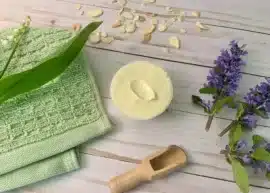 Almond body butter surrounded by purple flowers, lily of the valley, almonds, and a skincare scooper on a white wooden board.