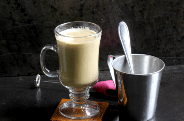 Starbucks flat white copycat in a glass mug with a handle, with a pewter cup and a spoon next to it, all on a black background with a pink flower