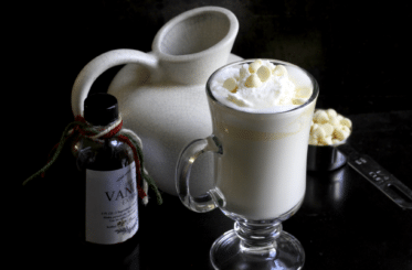 white hot chocolate with marshmallows on top in a glass mug with a small bottle of vanilla extract next to it and a white ceramic pitcher behind