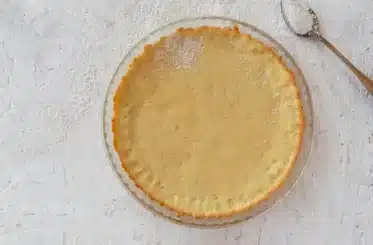 Coconut flour pie crust in a pie dish as seen from above, with a spoon next to it, all on a white surface