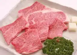 A plate of sliced Kobe beef accompanied by cubes of tallow beef and green vegetables.