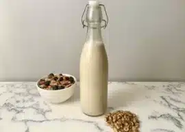 Homemade walnut milk in a glass jar on a marble countertop with a bowl of cereal to the left and walnuts to the right