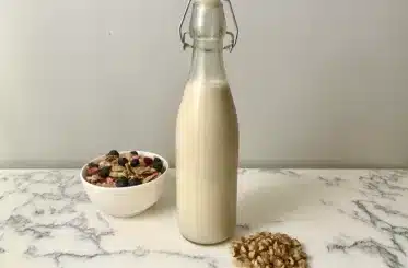 Homemade walnut milk in a glass jar on a marble countertop with a bowl of cereal to the left and walnuts to the right