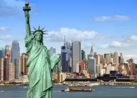 statue of liberty with new york city skyline behind and the hudson river in between, with a barge going across