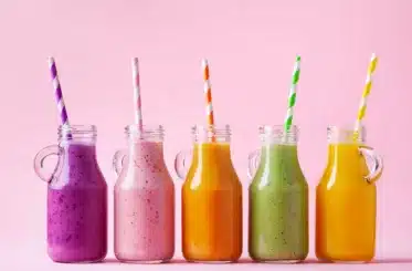 purple, pink, orange, green, and yellow smoothies in a line of glass bottles, each with a striped straw matching the color of the smoothie. All on a pink background