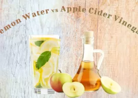 A wooden background showing a glass of lemon water and jug of apple cider vinegar accompanied by a few apples.