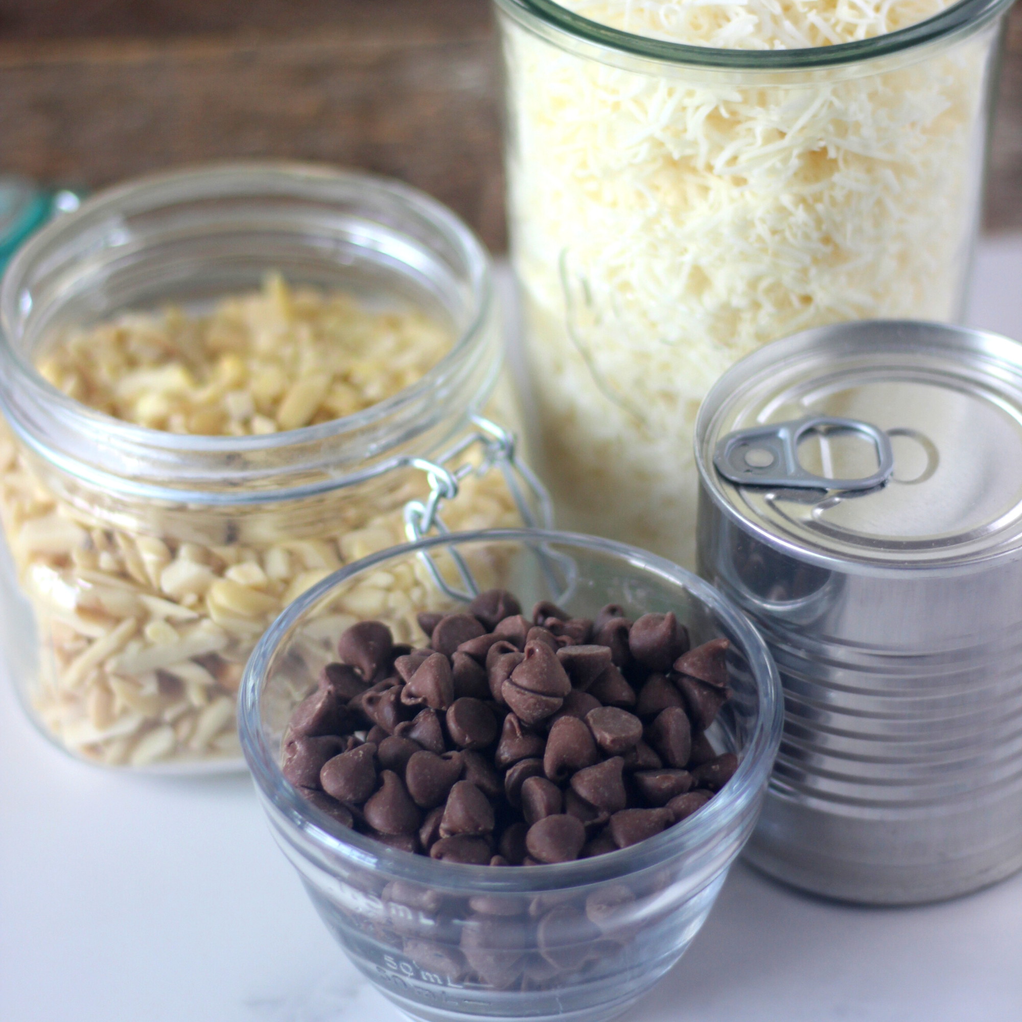 A large jar of shredded coconut, slivered almonds, chocolate chips and a tin of condensed milk. 
