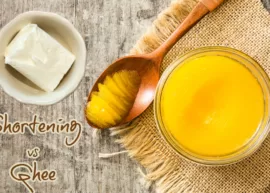 Aerial view of a wooden table set with a block of shortening in a bowl along with a bowl and a spoonful of ghee.