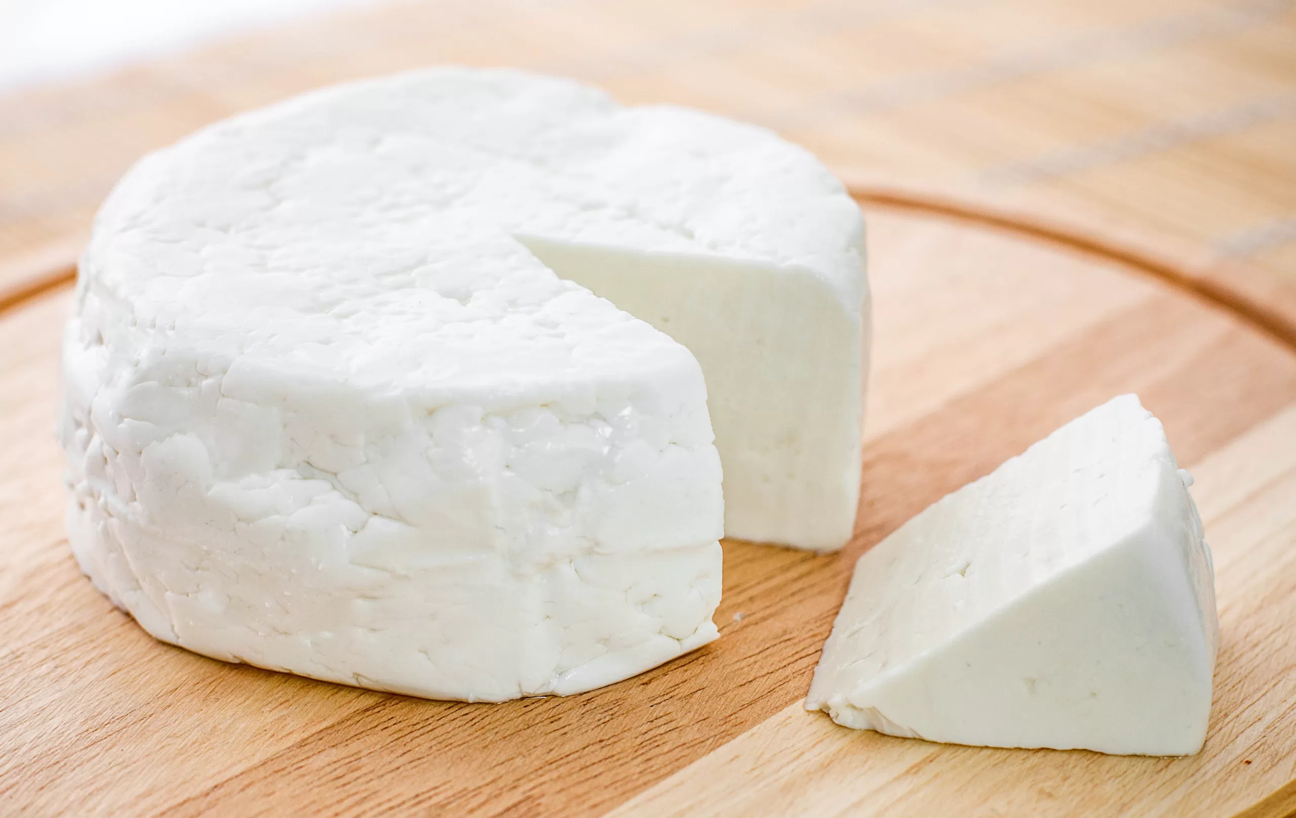 A round white cheese wheel with a wedge cut out on a wooden board.