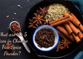 An aerial shot of a table featuring star anise, fennel seeds, szechuan peppercorns, whole cloves, and cinnamon stick. The text reads, "What are the 5 spices in Chinese Five Spice Powder?"