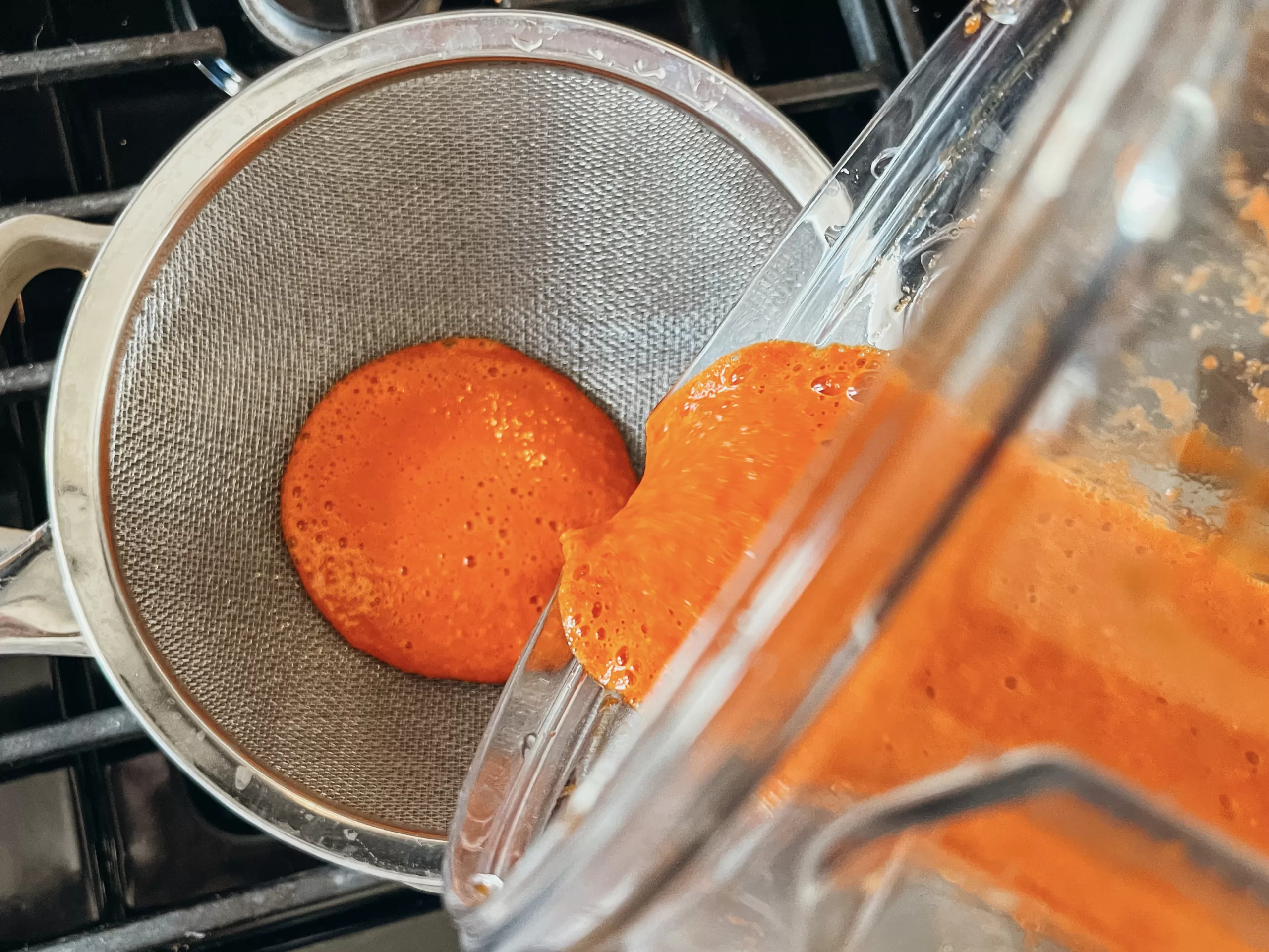 A blend of homemade sriracha sauce being poured from a blender, through a strainer, into a saucepan.