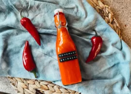 A bottle of homemade sriracha sauce on top of a blue towel surrounded by red jalapeño peppers.