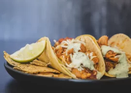 Three chicken tacos with sauce and lime wedge on a plate with tortilla chips.