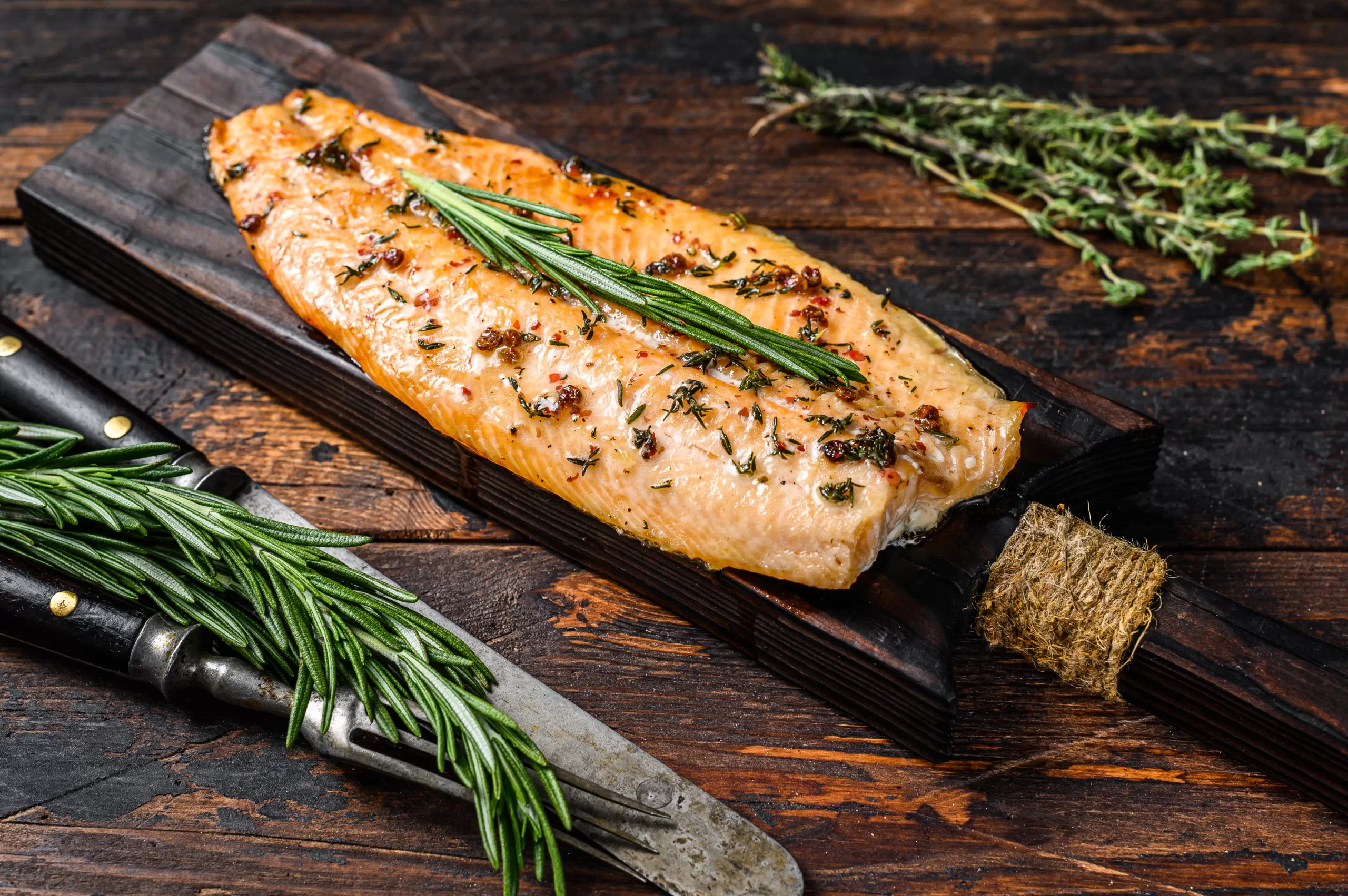 A wooden board holding a grilled trout fillet with thyme and rosemary.