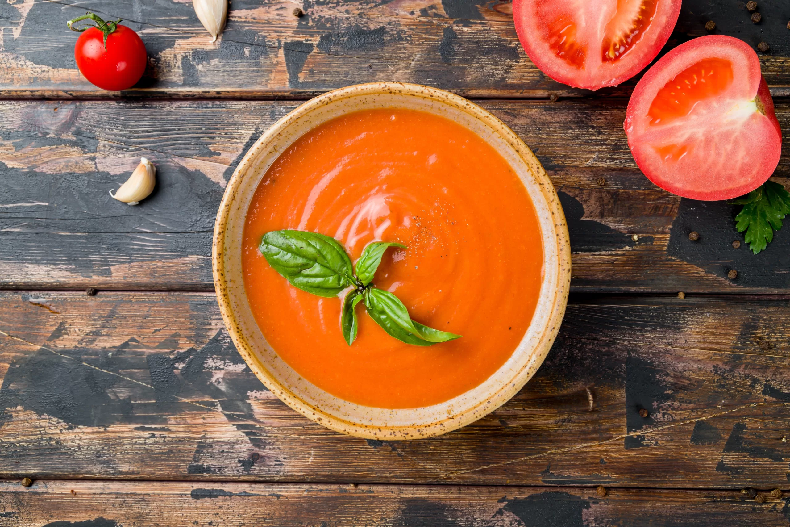 Top view of a vintage wooden table holding a bowl of tomato bisque and a few fresh tomatoes.