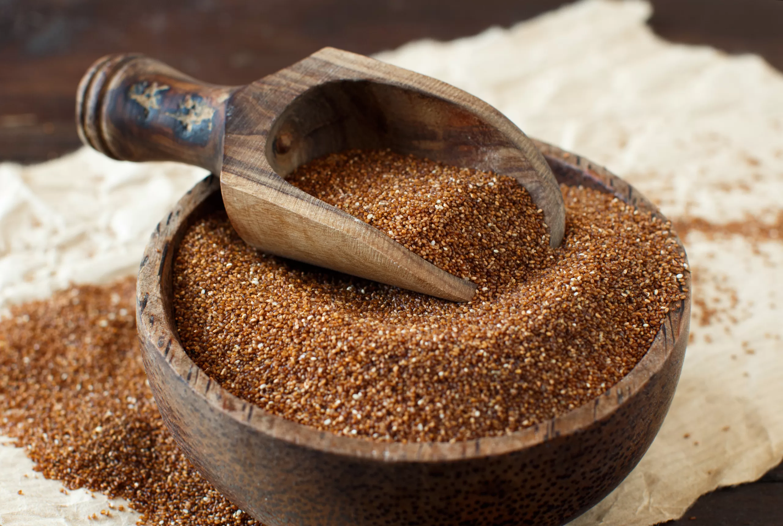 A bowl overflowing with raw teff and a measuring spoon on top.