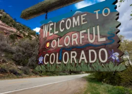 Sign that says "Welcome to Colorful Colorado" with a picture of mountains, grass, and a stream behind the text. The sign is suspended by a chain from a wooden post, and behind it is blue sky with some clouds, trees, and a steep mountain