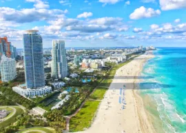 Miami Florida durnig the day - skyscrapers and a number of smaller buildings to the left, ocean to the right, beach in between, and blue sky with clouds above.