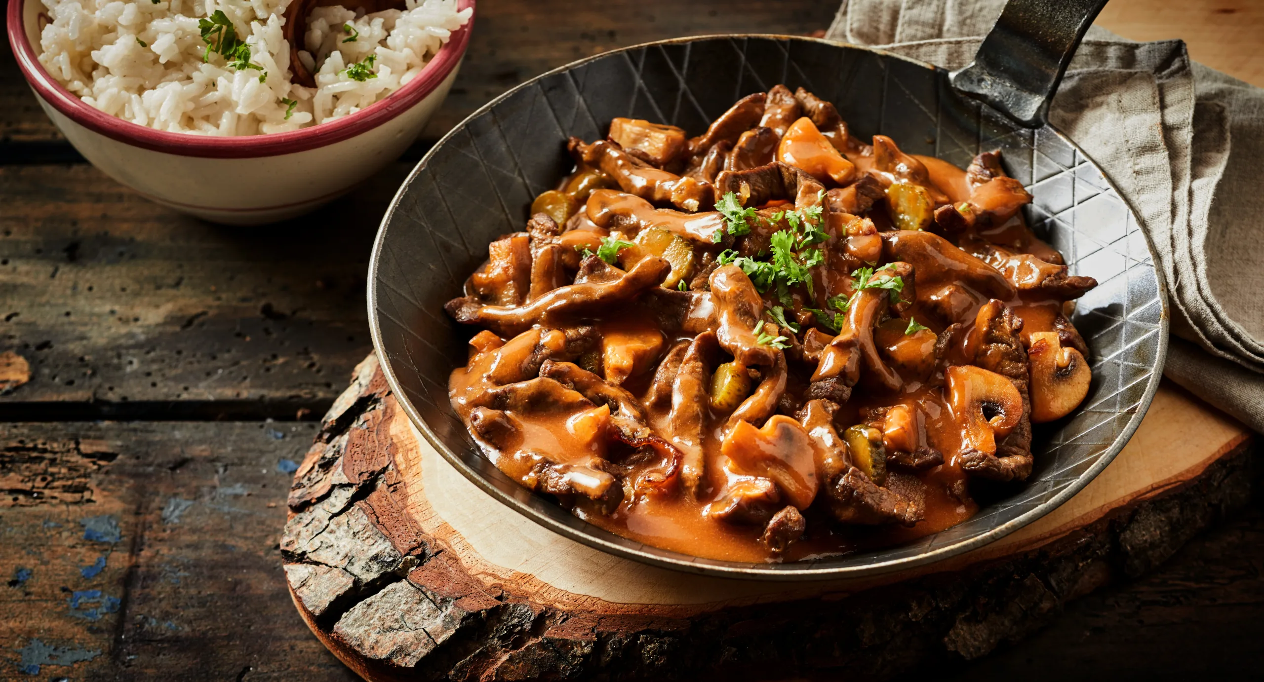 A rustic wooden surface displaying a frying pan with zürcher geschnetzeltes and a rice bowl. 