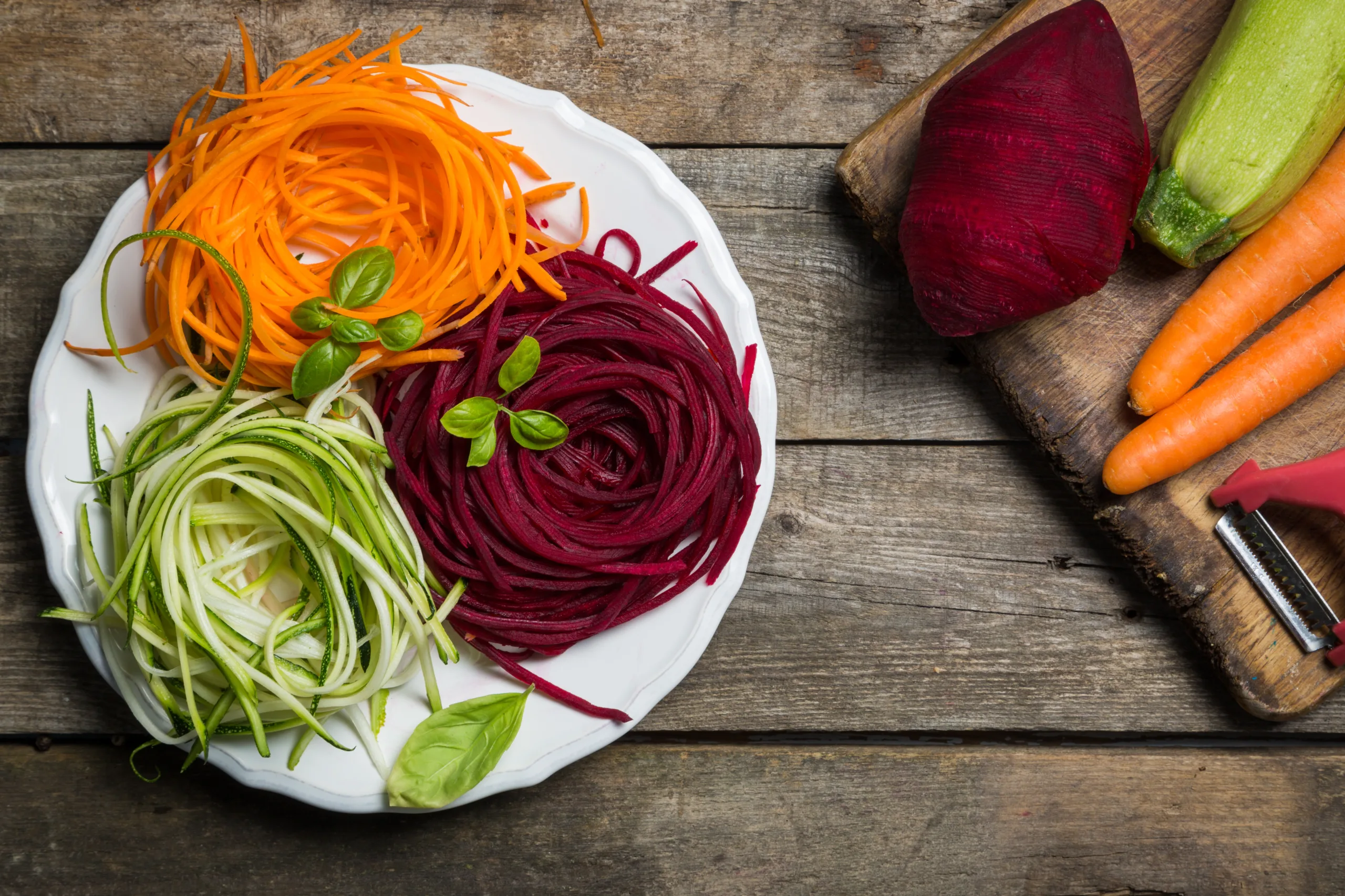 Top view of a rustic wooden table featuring a plate with carrot, zucchini, and beet zoodles.
