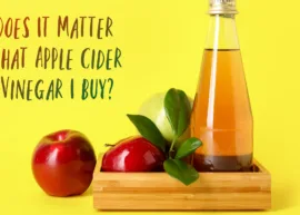 A bottle of apple cider vinegar encircled by some fresh apples against a yellow backdrop. The text reads, "Does It Matter What Apple Cider Vinegar I Buy?"