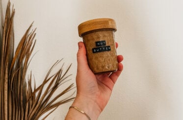 A hand holding a jar with a wood top and a label that states “Nut Butter.” A palm frond is to the left side.